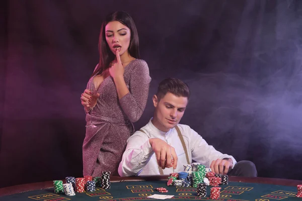 Hombre jugando al póquer en el casino, sentado en la mesa con montones de fichas y cartas en él. Celebrar la victoria con la mujer. Fondos negros de humo. Cierre. — Foto de Stock