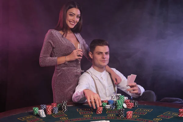 Man holding cards, playing poker at casino, sitting at table with stacks of chips on it. Celebrates win with woman. Black, smoke background. Close-up. — Stock Photo, Image