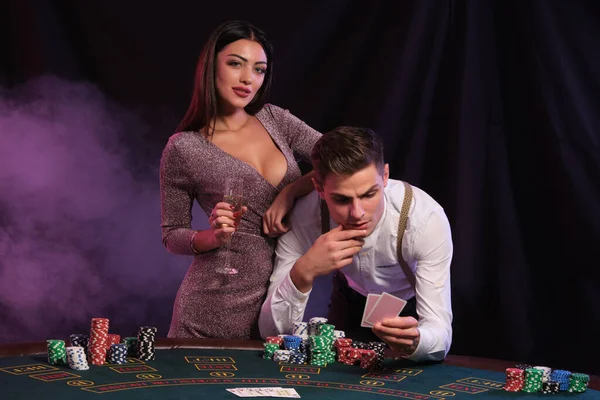 Hombre sosteniendo dos cartas, jugando al póquer en el casino, en la mesa con montones de fichas en ella. Celebra la victoria con la mujer. Fondos negros de humo. Cierre. — Foto de Stock