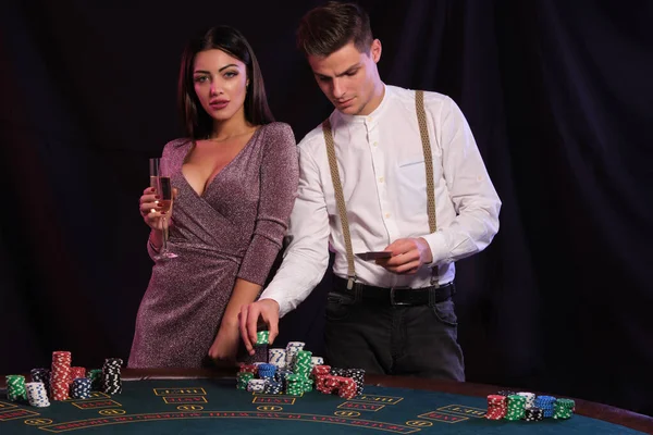 Hombre sosteniendo dos cartas, jugando al póquer en el casino, en la mesa con montones de fichas en ella. Celebra la victoria con una mujer sexy. Fondos negros. Cierre. — Foto de Stock