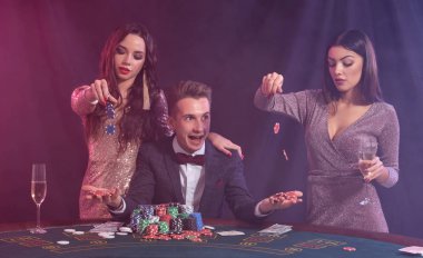 Kumarhanede poker oynayan adam masasında bir yığın fiş, para ve kartlarla oturuyor. İki kadınla zaferi kutluyoruz. Siyah, dumanlı arka plan. Yakın plan..