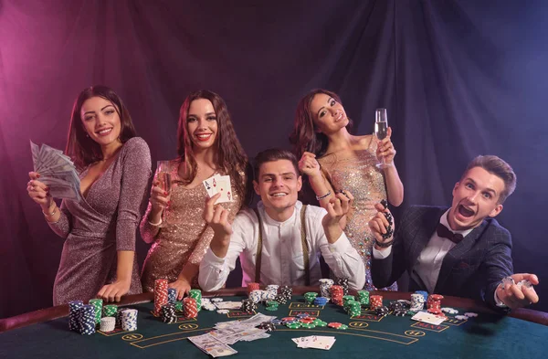 Amigos jugando al póquer en el casino, en la mesa con montones de fichas, dinero, cartas en él. Celebrar la victoria, sonriendo. Fondos negros de humo. Cierre. — Foto de Stock