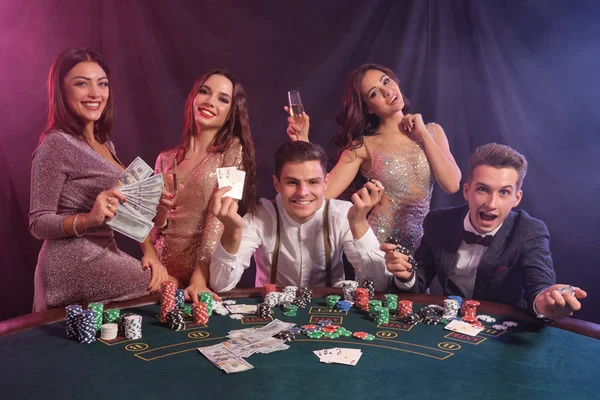 Amigos jugando al póquer en el casino, en la mesa con montones de fichas, dinero, cartas en él. Celebrar la victoria, sonriendo. Fondos negros de humo. Cierre. — Foto de Stock