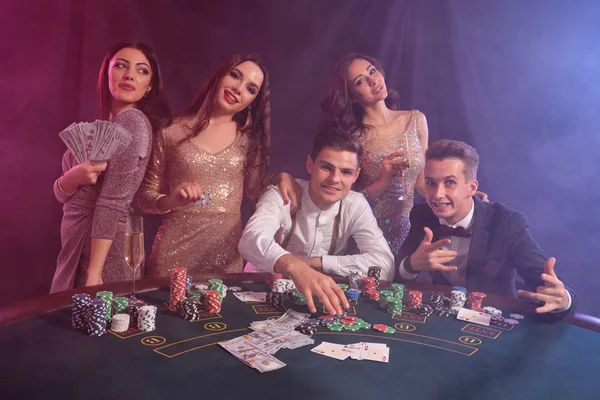 Kumarhanede poker oynayan arkadaşlar, masalarda yığınla fiş, para, kartlar. Zaferi kutluyor, gülümsüyor. Siyah, dumanlı arka plan. Yakın plan.. — Stok fotoğraf