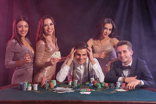 Přátelé hrají poker v kasinu, u stolu s hromadami žetonů, peněz, karet. Slavit vítězství, usmívat se. Černá, kouřové pozadí. Detailní záběr. — Stock fotografie