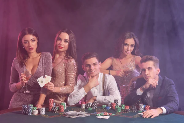 Amigos jugando al póquer en el casino, en la mesa con montones de fichas, dinero, cartas en él. Celebrar la victoria, sonriendo. Fondos negros de humo. Cierre. — Foto de Stock