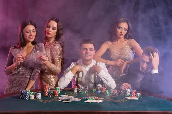 Vrienden die poker spelen in het casino, aan tafel met stapels chips, geld, kaarten erop. Winnen vieren, lachen. Zwart, rookachtergrond. Close-up. — Stockfoto