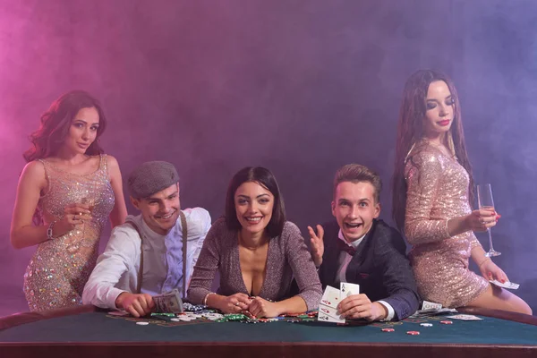 Přátelé hrají poker v kasinu, u stolu s hromadami žetonů, peněz, karet. Slavit vítězství, usmívat se. Černá, kouřové pozadí. Detailní záběr. — Stock fotografie