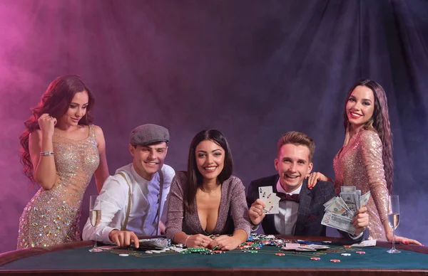 Amis jouant au poker au casino, à la table avec des piles de jetons, de l'argent, des cartes sur lui. Célébration de la victoire, sourire. Noir, fond fumigène. Gros plan. — Photo