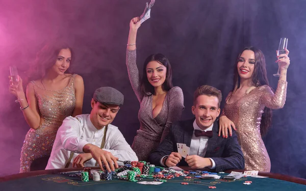Freunde spielen Poker im Casino, am Tisch mit Stapeln von Chips, Geld, Karten darauf. Sieg feiern, lächeln. Schwarzer, rauchiger Hintergrund. Nahaufnahme. — Stockfoto