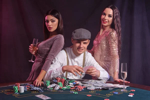 Hombre jugando al póquer en el casino sentado a la mesa con montones de fichas, dinero, cartas. Celebrar la victoria con dos mujeres. Fondos negros. Cierre. —  Fotos de Stock