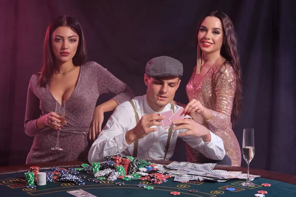 Man playing poker at casino sitting at table with stacks of chips, money, cards. Celebrating win with two women. Black background. Close-up. — Stock Photo, Image