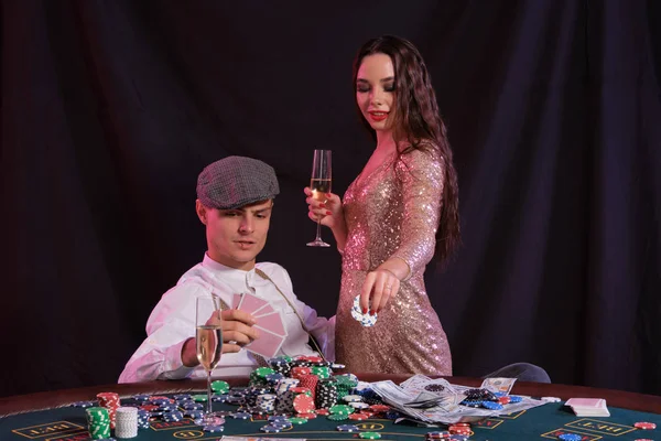 Homme jouant au poker au casino assis à table avec des piles de jetons, argent, champagne, cartes. Célébrer la victoire avec une femme. Fond noir. Gros plan . — Photo
