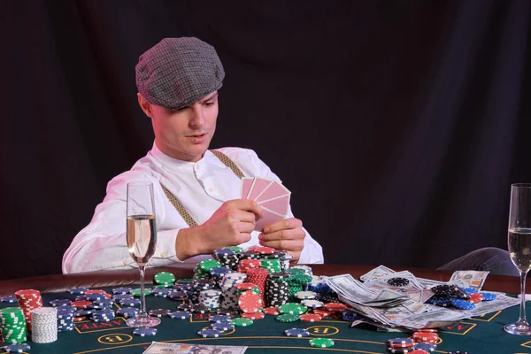 Mann, der im Casino Poker spielt, sitzt am Tisch und stapelt Chips, Geld, Champagner, Karten. Schwarzer Hintergrund, bunte Hintergrundbeleuchtung. Nahaufnahme. — Stockfoto