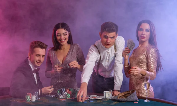 Amici che giocano a poker al casinò, al tavolo con pile di chips, soldi, carte su di esso. Celebrare vincere, sorridere. Nero, sfondo fumo. Primo piano. — Foto Stock