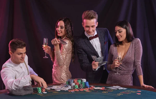 Amigos jugando al póquer en el casino, en la mesa con montones de fichas, dinero, cartas en él. Celebrar la victoria, sonriendo. Fondos negros. Cierre. — Foto de Stock