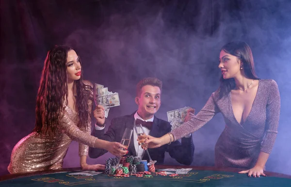 Mann, der im Casino Poker spielt, sitzt am Tisch und stapelt Chips, Geld und Karten. Mit zwei Frauen den Sieg feiern. Schwarzer, rauchiger Hintergrund. Nahaufnahme. — Stockfoto