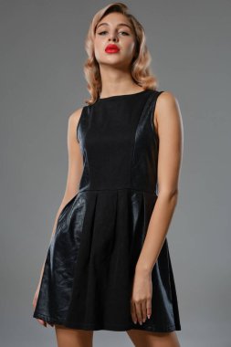 Siyah kısa deri elbiseli, parlak makyajlı güzel sarışın kız gri stüdyo arka planında poz veriyor. Moda ve güzellik. Yakın plan..