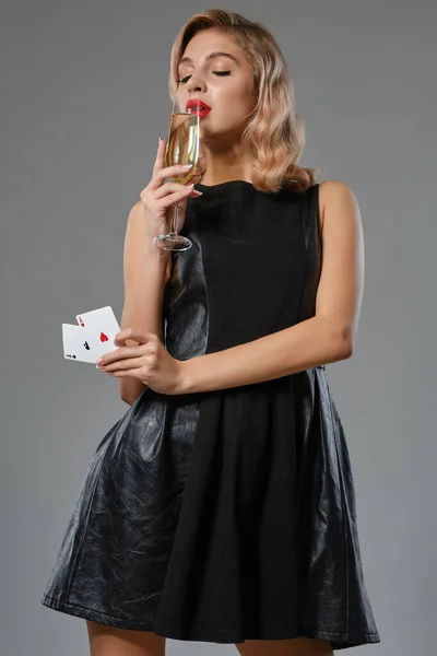 Blonde girl in black dress showing two playing cards, drinking champagne, posing against gray studio background. Gambling, poker, casino. Close-up. — Stock Photo, Image