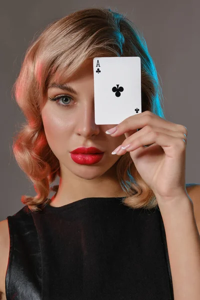 Blondýnka v černých kožených šatech ukazuje eso z holí, pózuje na šedém pozadí. Hazardní hry, poker, kasino. Detailní záběr. — Stock fotografie