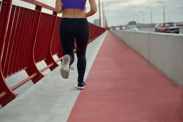 Дівчина-спортсмен біжить вранці на вулиці, в місті, на сучасному мосту, що перетинає річку. Активний здоровий спосіб життя. Вид ззаду . — стокове фото