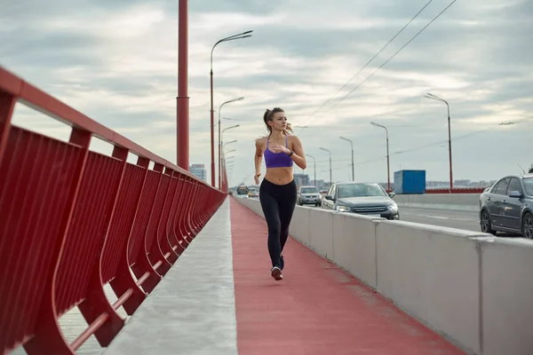 Atleta menina está correndo pela manhã fora, na cidade, na ponte moderna que cruza o rio. Estilo de vida saudável ativo. Conceito de esporte e recreação. — Fotografia de Stock