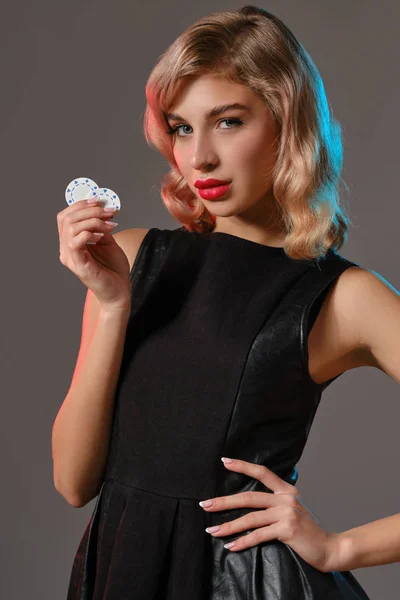 Blond meisje in zwarte stijlvolle jurk met twee chips, poseren tegen grijze achtergrond. Gokken entertainment, poker, casino. Close-up. — Stockfoto