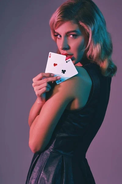 Девушка-блондинка в черном стильном платье показывает две игральные карты, позируя на цветном фоне. Азартные игры, покер, казино. Закрытие. — стоковое фото