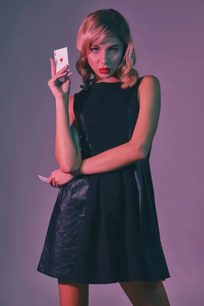 Девушка-блондинка в черном стильном платье показывает две игральные карты, позируя на цветном фоне. Азартные игры, покер, казино. Закрытие. — стоковое фото