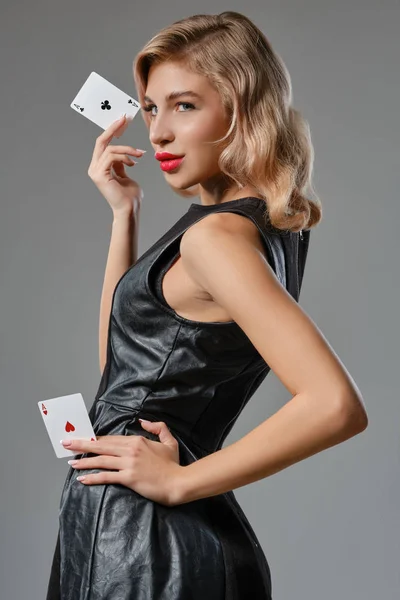 Blondýny dívka v černých stylových šatech ukazující dvě hrací karty, pózující na šedém pozadí. Hazardní hry, poker, kasino. Detailní záběr. — Stock fotografie