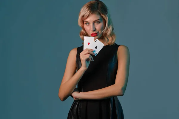 Chica rubia en vestido elegante negro que muestra dos cartas de juego, posando sobre fondo azul. Entretenimiento, póquer, casino. Primer plano . —  Fotos de Stock