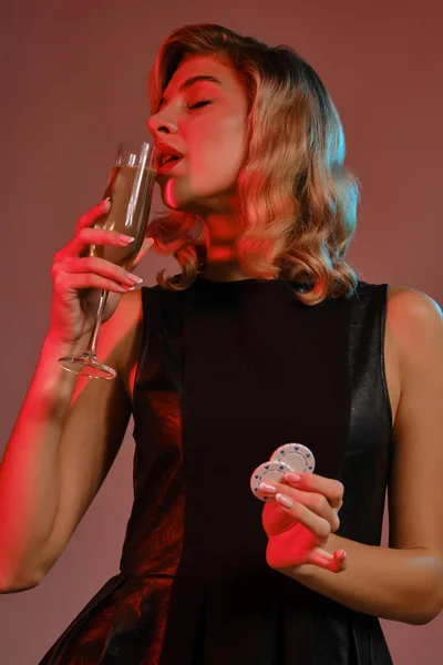 Девушка-блондинка в черном платье держит в руках чипсы и пьет шампанское, позируя на цветном фоне. Азартные игры, покер, казино. Закрытие. — стоковое фото