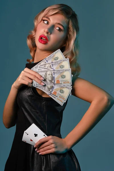 Menina loira em vestido elegante preto segurando algum dinheiro e cartões, posando contra fundo azul. Jogos de entretenimento, poker, cassino. Close-up. — Fotografia de Stock