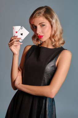 Siyah şık elbiseli sarışın kız gri arka planda poz veren iki iskambil kağıdı gösteriyor. Kumar eğlencesi, poker, kumarhane. Yakın plan..