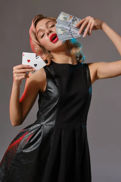 黒のスタイリッシュなドレスのブロンドの女の子は、灰色の背景に対してポーズをいくつかのお金とカードを保持。ギャンブルエンターテイメント,ポーカー,カジノ.閉鎖. — ストック写真