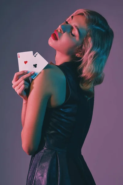 Siyah şık elbiseli sarışın kız renkli arka planda poz veren iki iskambil kağıdı gösteriyor. Kumar eğlencesi, poker, kumarhane. Yakın plan.. — Stok fotoğraf