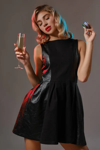 Ragazza bionda in abito nero in possesso di vetro di champagne e patatine fritte, posa su sfondo grigio. Gioco d'azzardo, poker, casinò. Primo piano. — Foto Stock