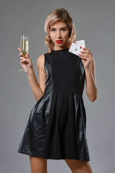 Blondes Mädchen in schwarzem Kleid, zwei Spielkarten und ein Glas Champagner in der Hand, posierend vor grauem Hintergrund. Glücksspiel, Poker, Casino. Nahaufnahme. — Stockfoto