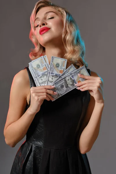 黒のスタイリッシュなドレスのブロンドの女の子は、灰色の背景に対してポーズをいくつかのお金を保持。ギャンブルエンターテイメント,ポーカー,カジノ.閉鎖. — ストック写真