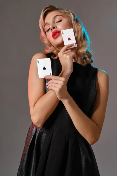 黒のスタイリッシュなドレスのブロンドの女の子は、灰色の背景に対してポーズを2枚のトランプを示しています。ギャンブルエンターテイメント,ポーカー,カジノ.閉鎖. — ストック写真