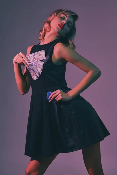 Ξανθιά κοπέλα σε μαύρο κομψό φόρεμα κρατώντας κάποια χρήματα και μάρκες, θέτοντας σε πολύχρωμο φόντο. Ψυχαγωγία, πόκερ, καζίνο. Κοντινό πλάνο. — Φωτογραφία Αρχείου