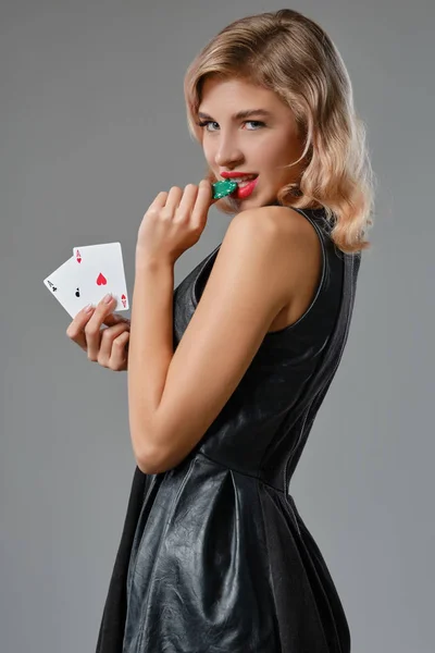 黒のドレスのブロンドの女の子は、横に灰色の背景に対してポーズを2枚のトランプと緑のチップを保持。ギャンブル、ポーカー、カジノ。閉鎖. — ストック写真