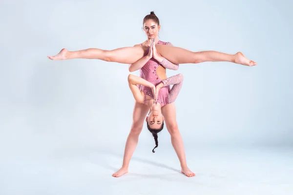 Två flexibla flickor gymnaster med flätor, i rosa trikåer utför splittringar med hjälp av stöd samtidigt poserar isolerad på vit bakgrund. Närbild. — Stockfoto