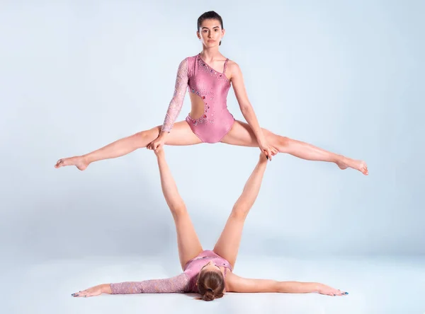 Δύο ευέλικτα κορίτσια γυμναστές με κοτσίδες, σε ροζ λεοπαρδάλεις εκτελούν σχισμές χρησιμοποιώντας υποστήριξη, ενώ θέτουν απομονώνονται σε λευκό φόντο. Κοντινό πλάνο. — Φωτογραφία Αρχείου