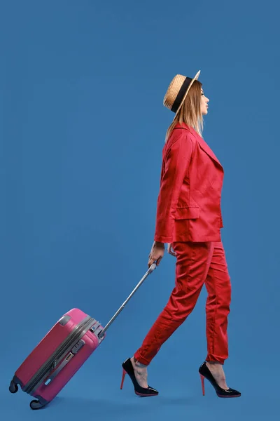 Blondýnka v klobouku, červený kalhotový kostým, vysoké podpatky. Nosí růžový kufr po rukojeti, kráčí po modrém pozadí. Boční pohled, plná délka — Stock fotografie