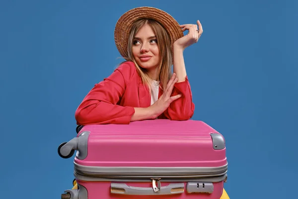 Blondýnka v slamáku, bílá blůza, červená bunda. Sedí opřený o barevné kufry, pózuje na modrém pozadí. Koncept cestování. Detailní záběr — Stock fotografie