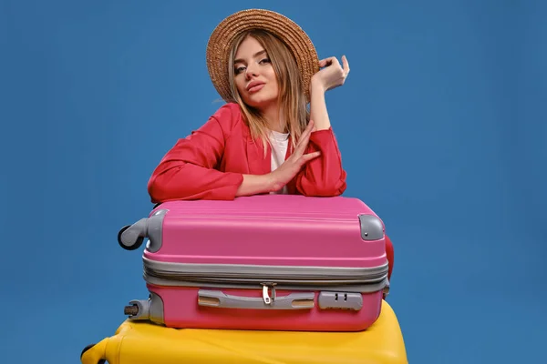 Donna bionda con cappello di paglia, camicetta bianca, giacca rossa. Seduto appoggiato su valigie colorate, in posa su sfondo blu. Concetto di viaggio. Primo piano — Foto Stock