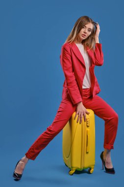 Beyaz bluzlu sarışın, kırmızı takım elbiseli, yüksek siyah topuklu. Saça dokunuyor, sarı bavulun üzerinde oturuyor, mavi arka planda poz veriyor. Tam uzunluk