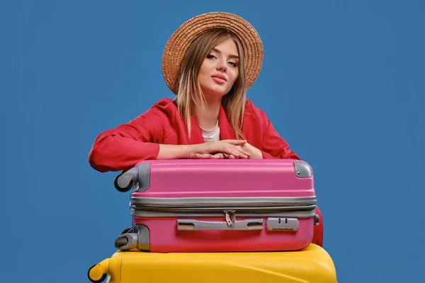 Блондинка в соломенной шляпе, белая блузка, красная куртка. Сидя, опираясь на красочные чемоданы, позируя на синем фоне. Концепция путешествия. Крупный план — стоковое фото