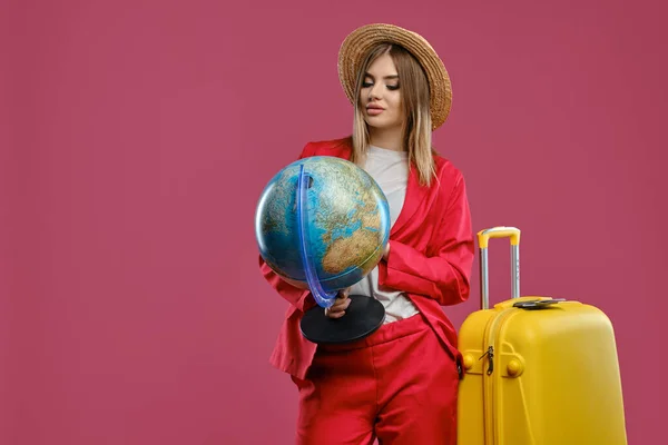 Модель в шляпе, белая блузка, красный брючный костюм. Она держит глобус, позирует на розовом фоне. Рядом есть желтый чемодан с паспортом и билетом. Крупный план — стоковое фото